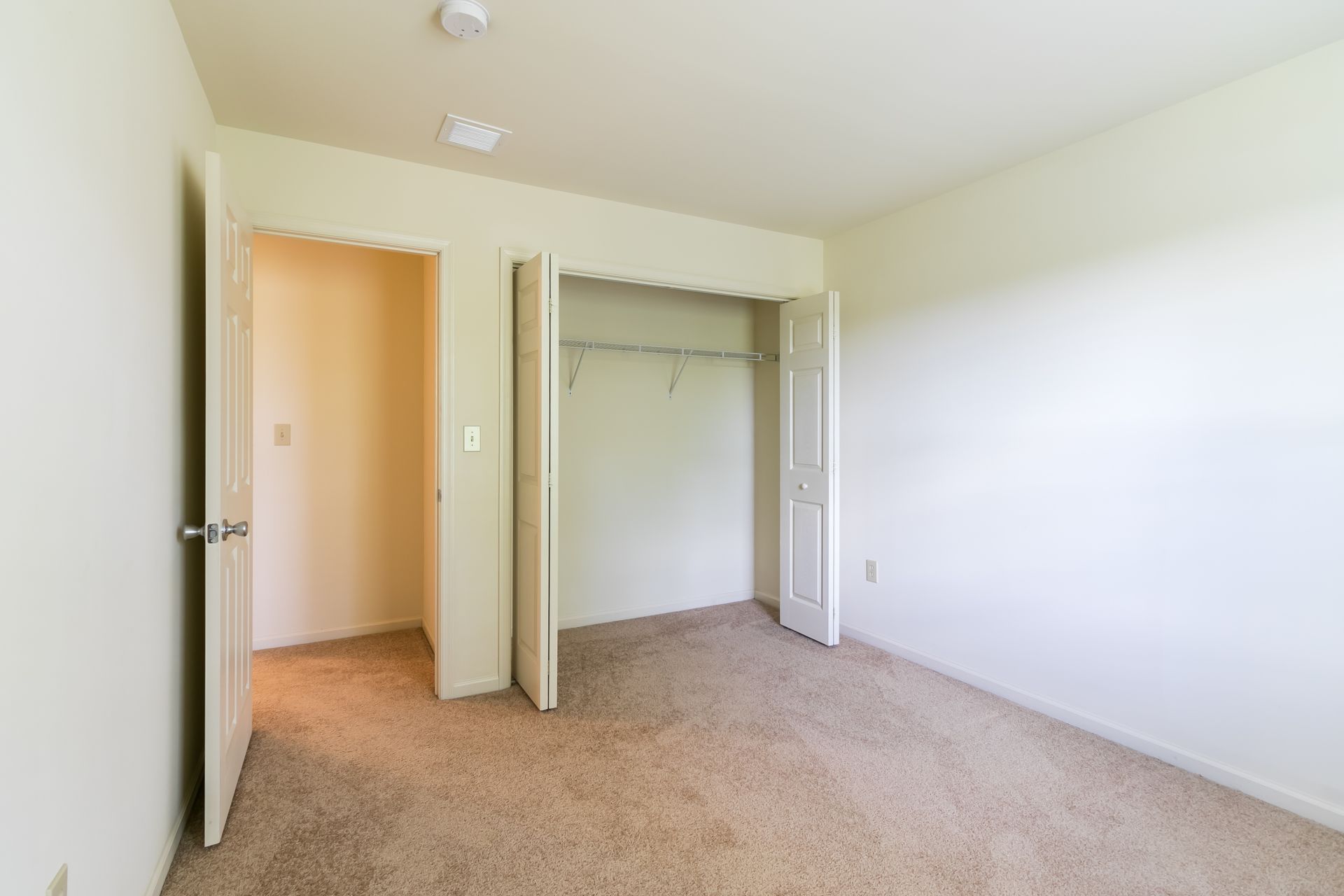 Empty beige bedroom with a closet and an open doorway; tan carpet.