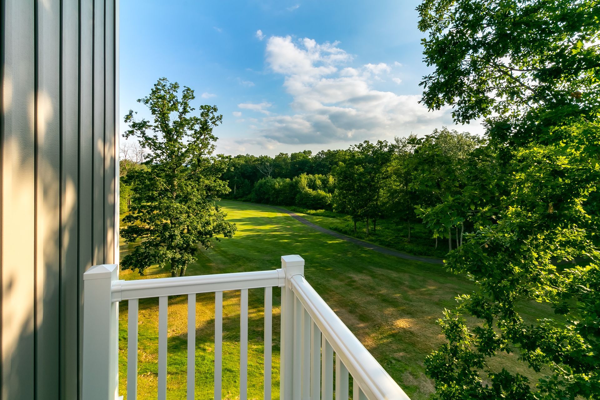 Balcony overlooking a green field and trees under a blue sky with clouds.