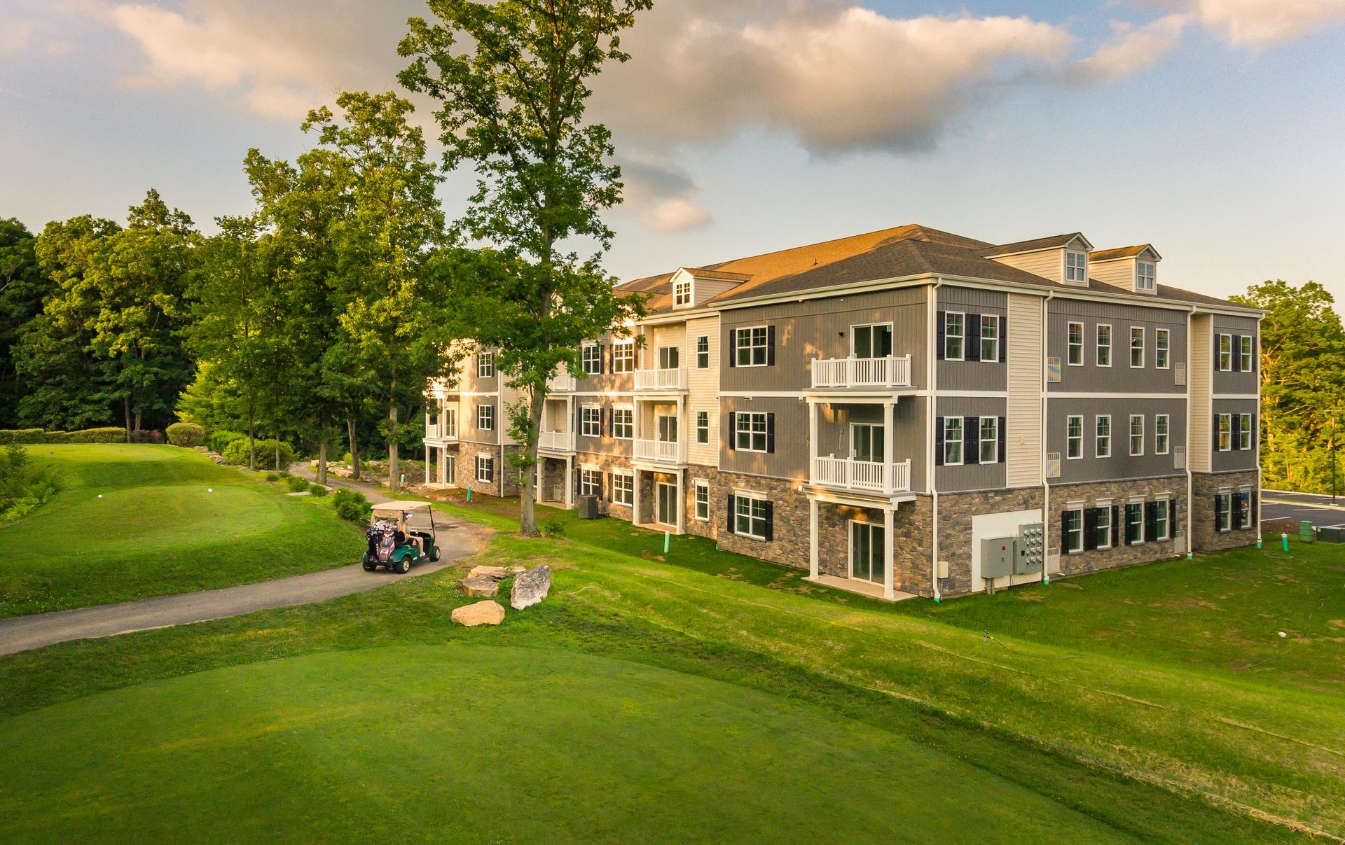 Apartment building next to a golf course. A golf cart is on the green. Green grass, trees, and gray building.