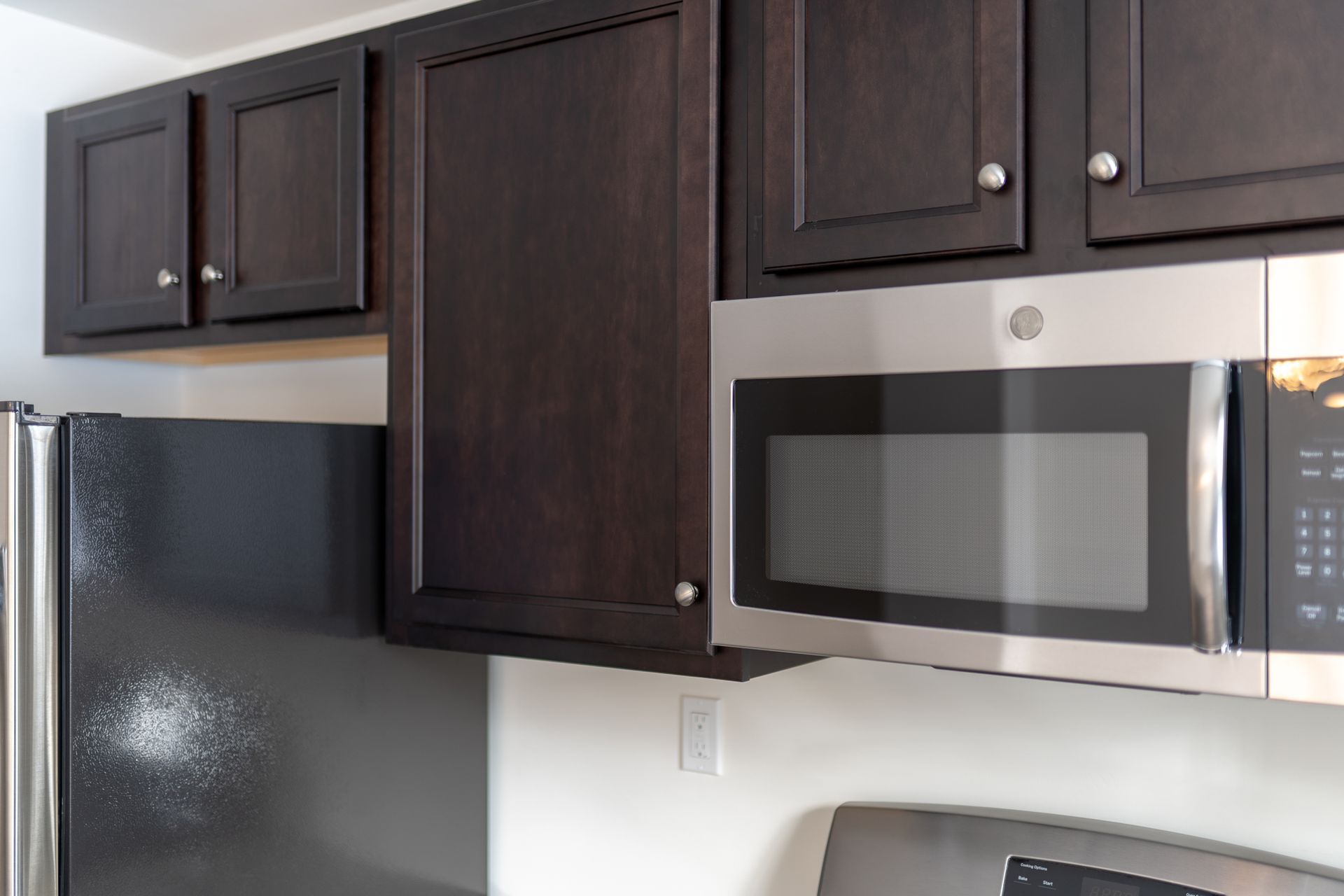 Dark wood kitchen cabinets above a stainless steel microwave and refrigerator. White walls.