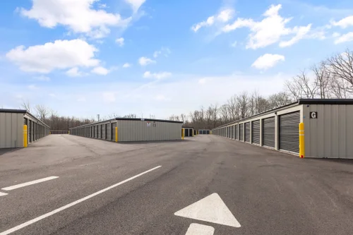 Rows of gray storage units with black doors and an asphalt driveway under a blue sky.