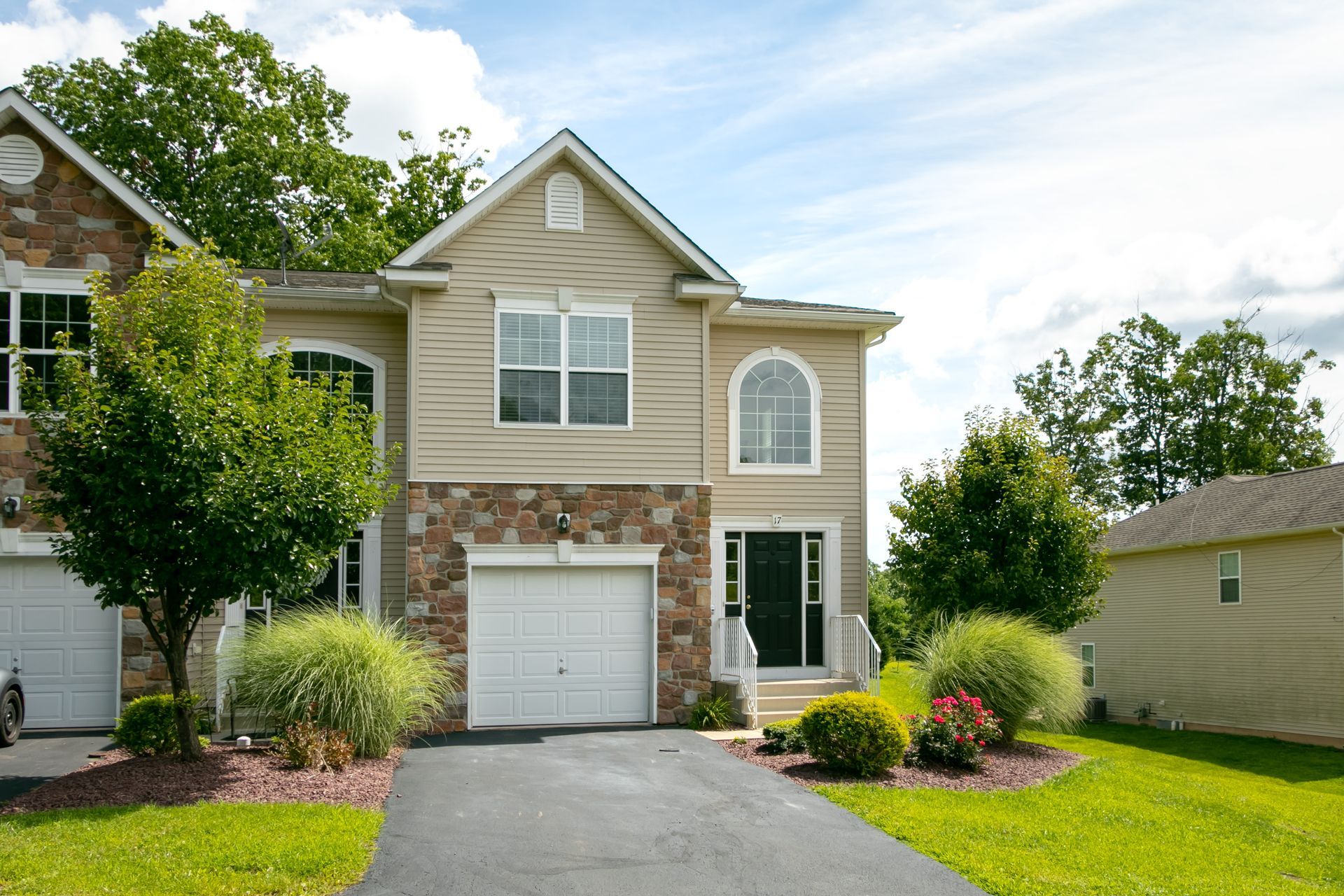 Two-story tan and stone townhouse with a garage, front door, and green lawn under a blue sky.