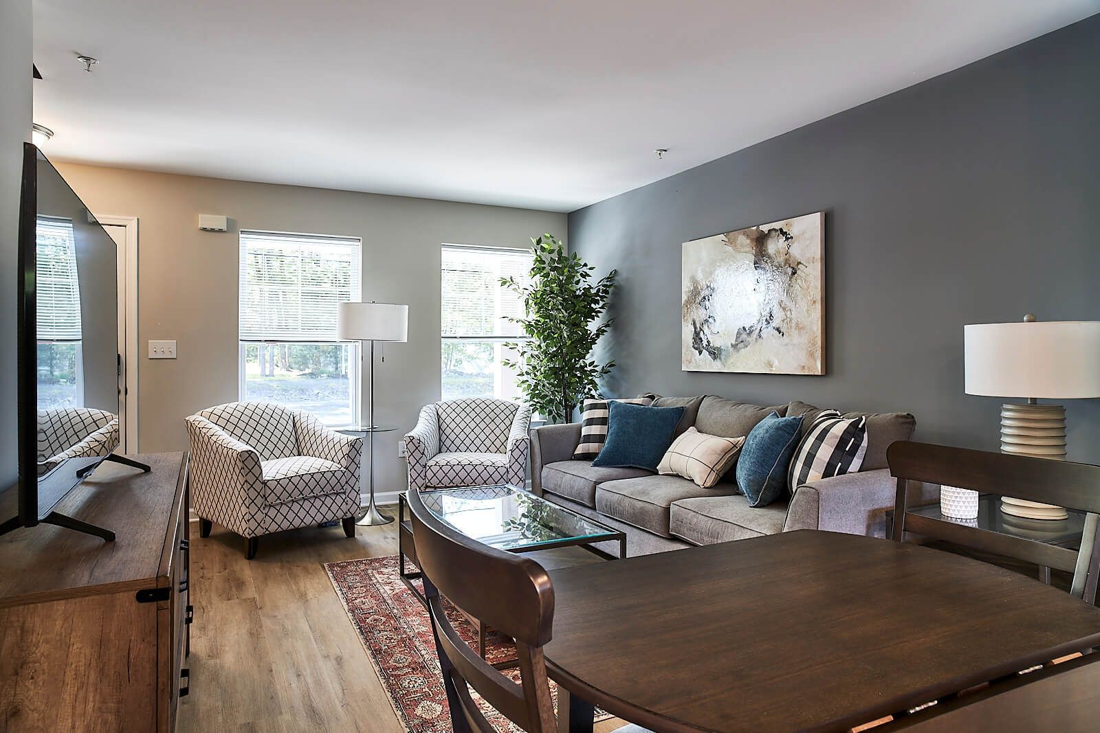 Living room with sofa, chairs, table, large TV, and art on the wall. Light grey walls, wooden floor.