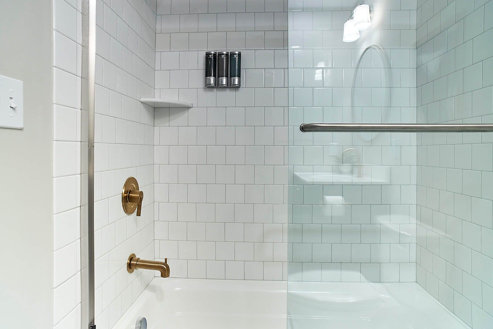 White tiled bathroom with glass shower door, gold fixtures, and soap dispensers.