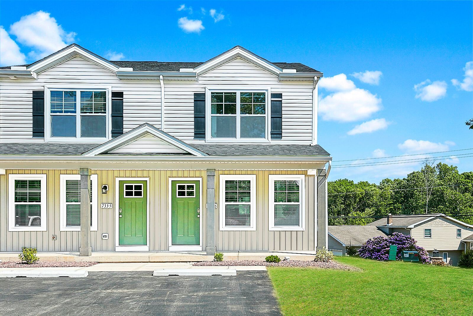 Two-story townhouses with light siding and green front doors under a blue sky, lush green lawn.