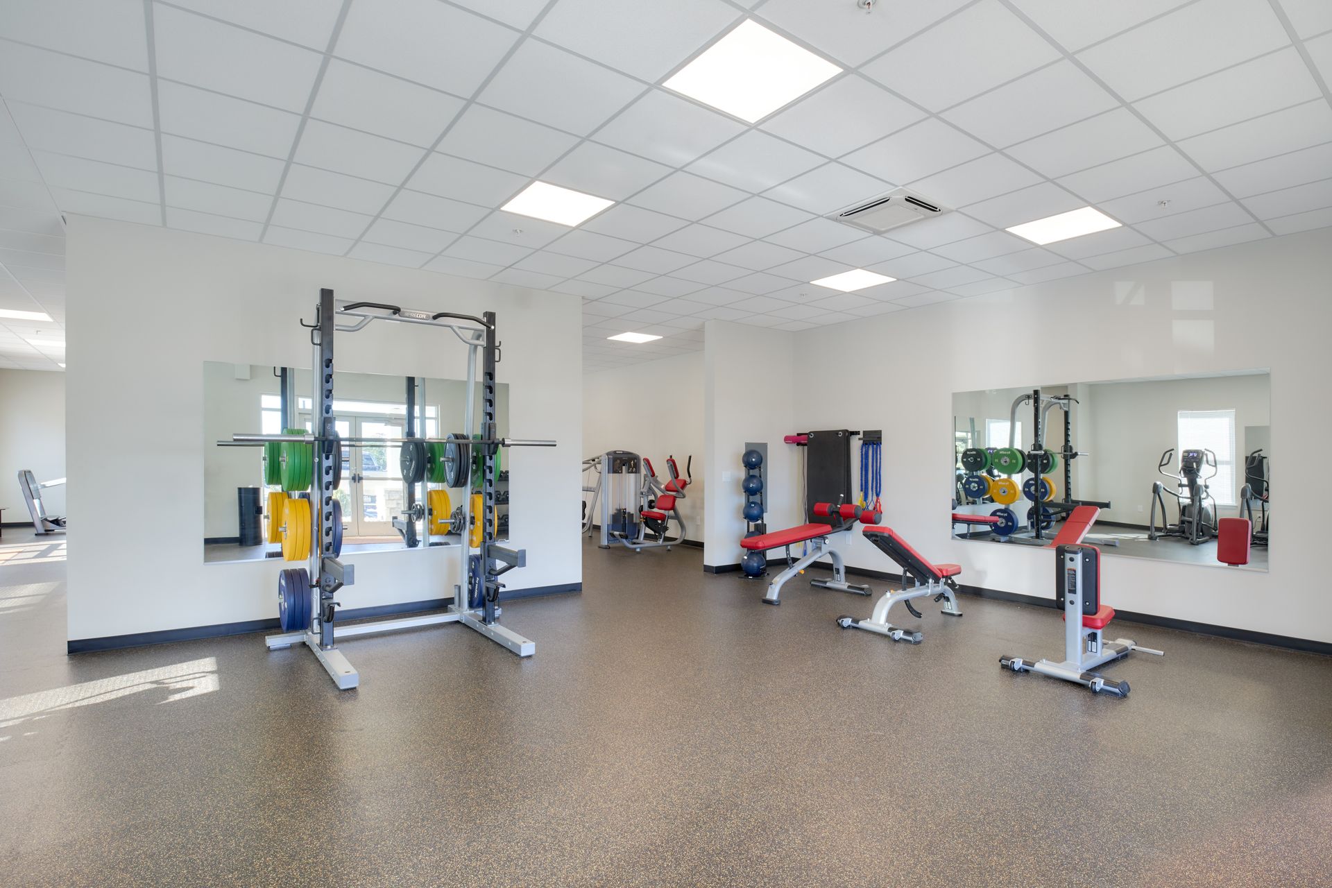 Gym with weightlifting equipment, mirrors, and exercise machines on a speckled floor.