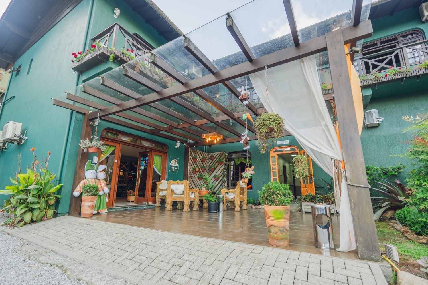 Edifício verde com entrada coberta, móveis de madeira e plantas em vasos.