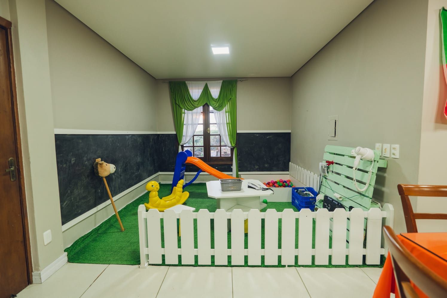 Sala de jogos interna com cerca branca, escorregador e brinquedos em grama artificial.