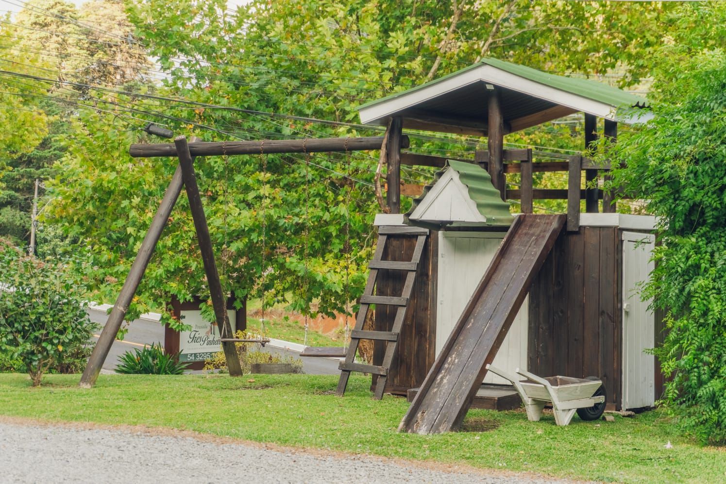 Parque infantil com balanço, escorregador e pequena estrutura de madeira em área gramada, cercada por árvores.