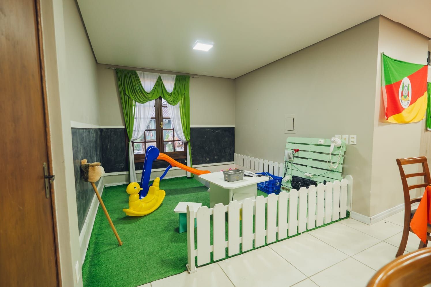 Área de recreação infantil interna com gangorra, cerca branca, grama sintética e uma bandeira brasileira.