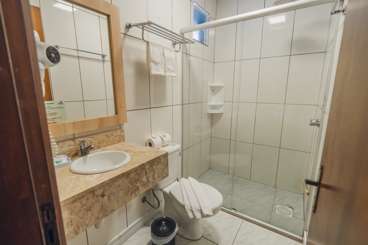 Banheiro com chuveiro, vaso sanitário, pia e espelho. Azulejos brancos e bege.