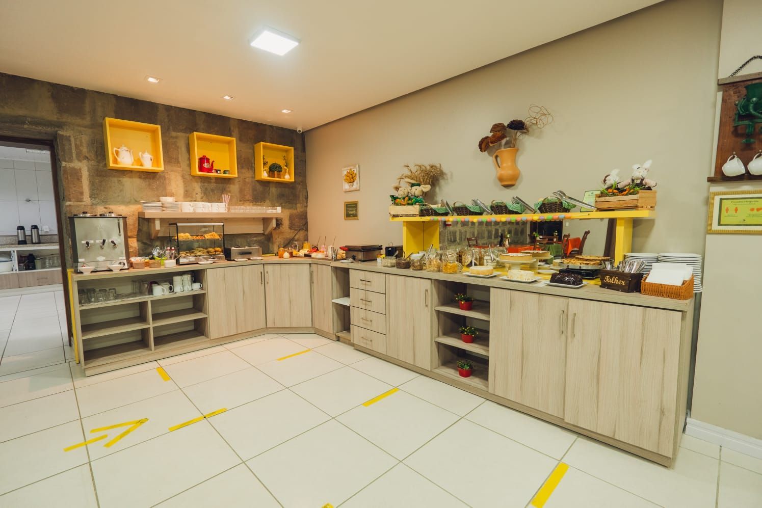 Buffet de café da manhã: balcão de alimentos com vários pratos, detalhes em amarelo e marcações no chão também em amarelo.