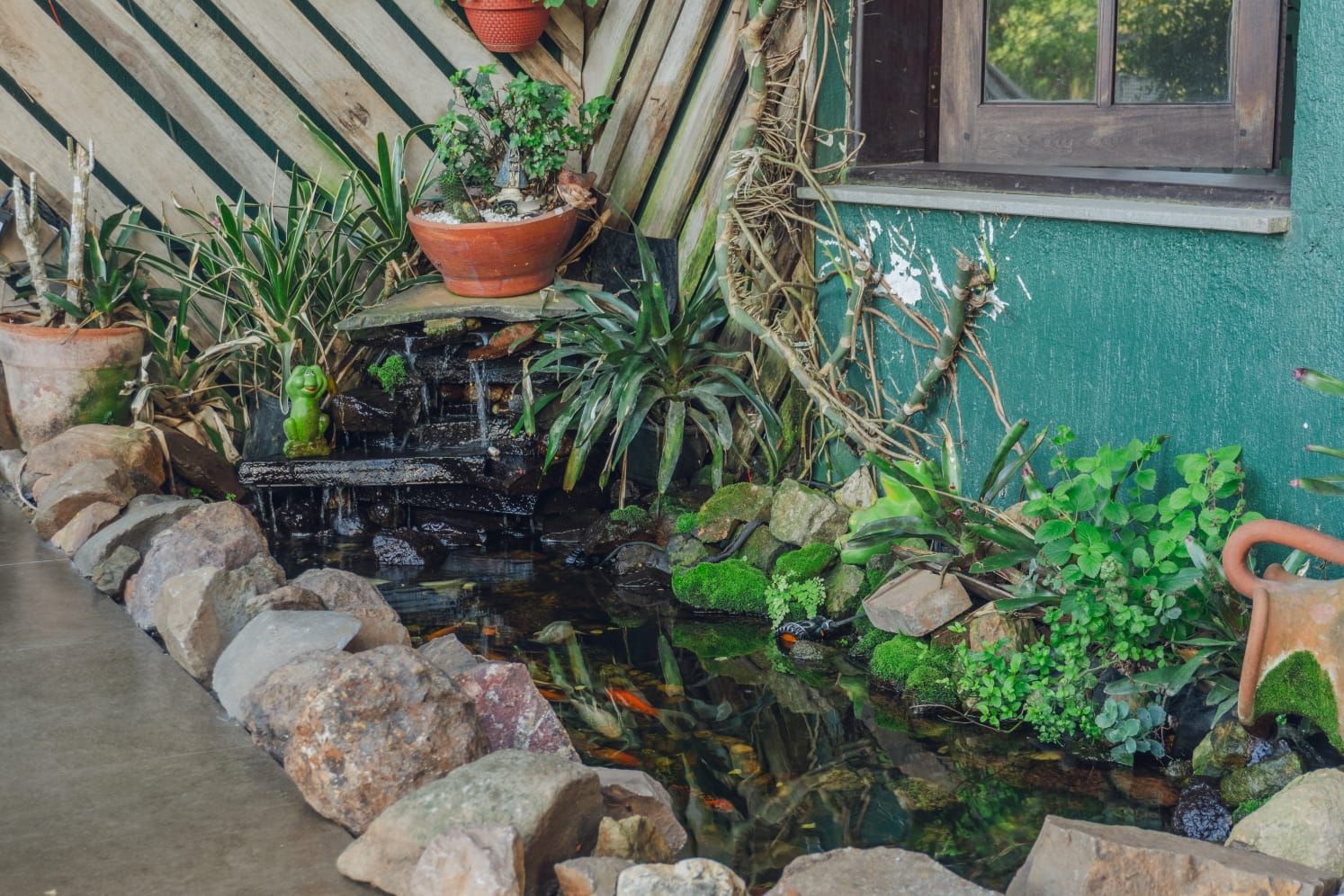 Um pequeno lago ao ar livre com uma cachoeira, cercado por pedras e vegetação exuberante.