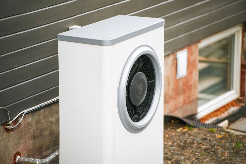 A white air conditioner is sitting outside of a house next to a window.