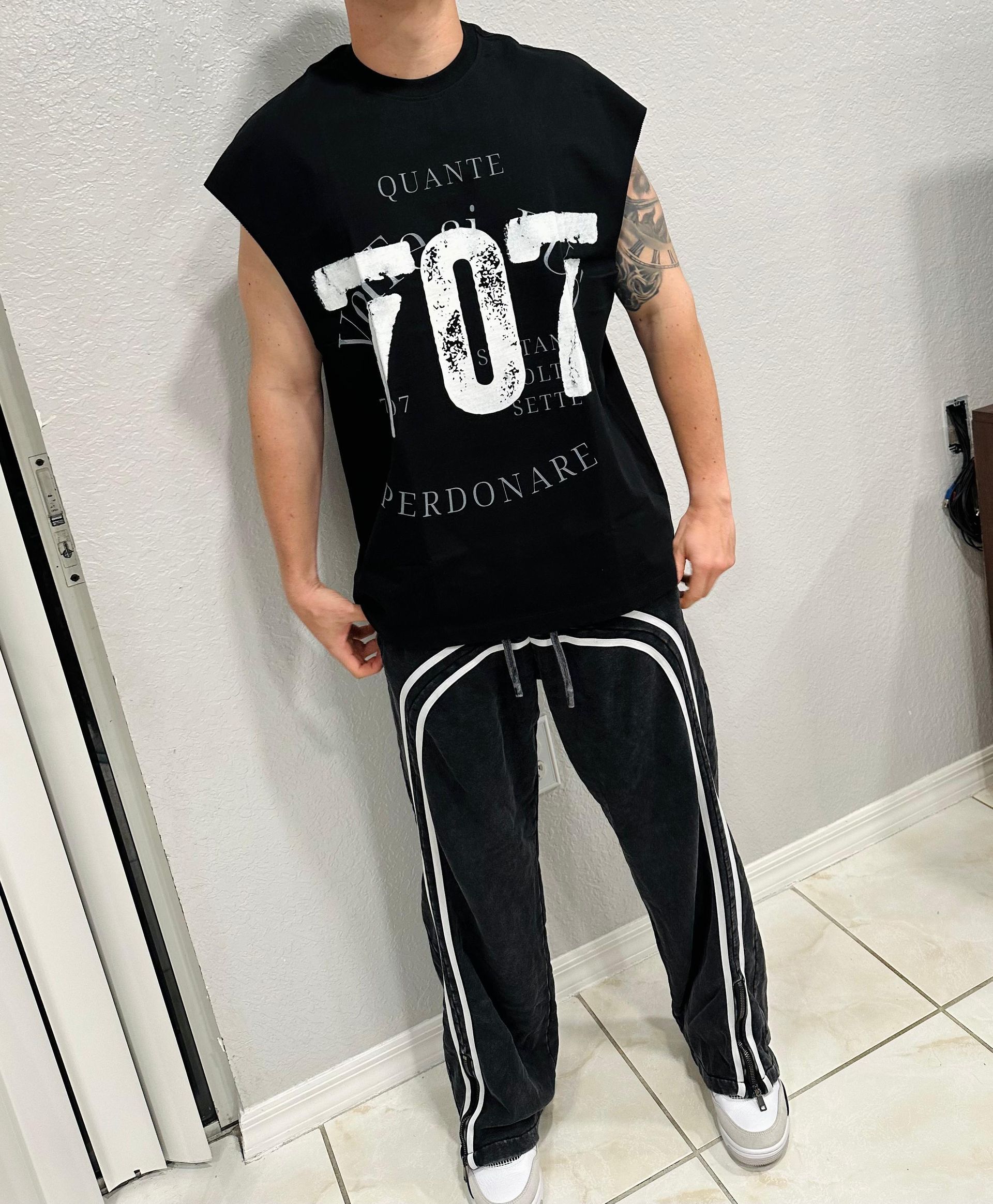 Man in black sleeveless shirt and track pants with white stripes, leaning against a wall.