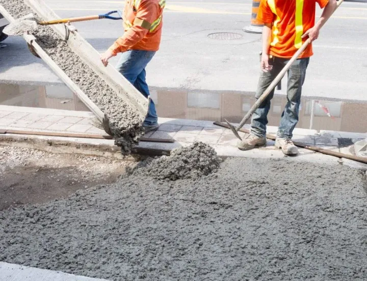 Two workers in bright orange safety shirts pouring wet concrete from a truck chute onto a construction site.