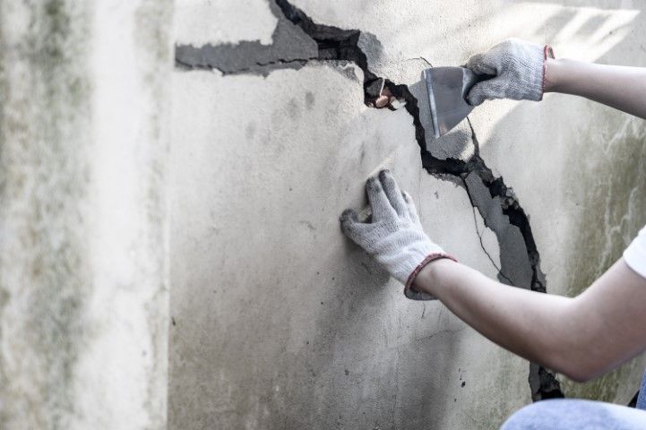 A person wearing work gloves uses a putty knife to repair a large crack in a concrete wall.