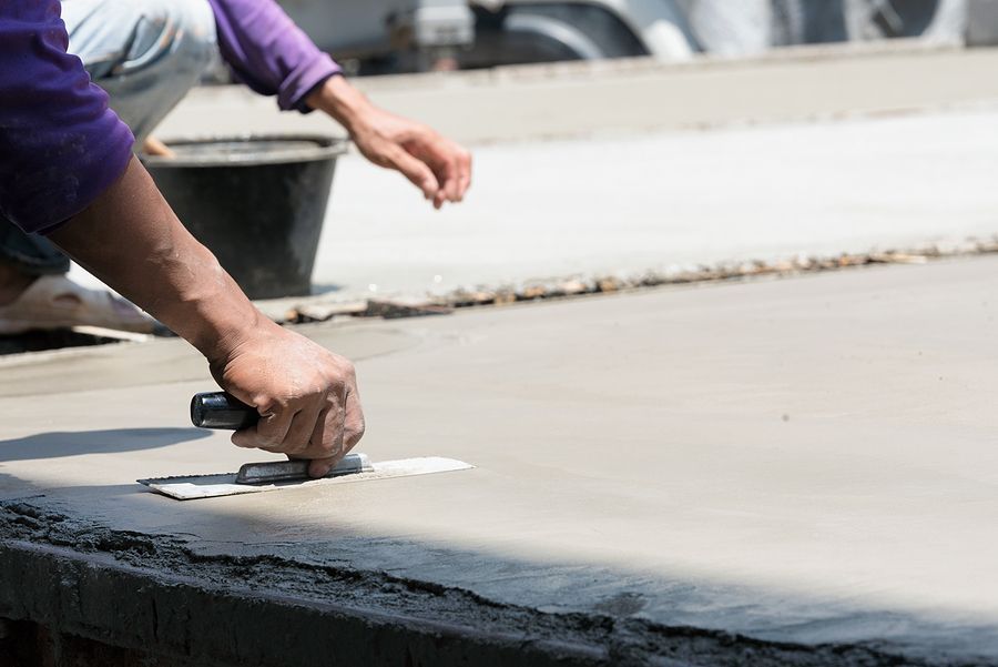 A person in a purple shirt uses a metal trowel to smooth wet concrete on a construction site.