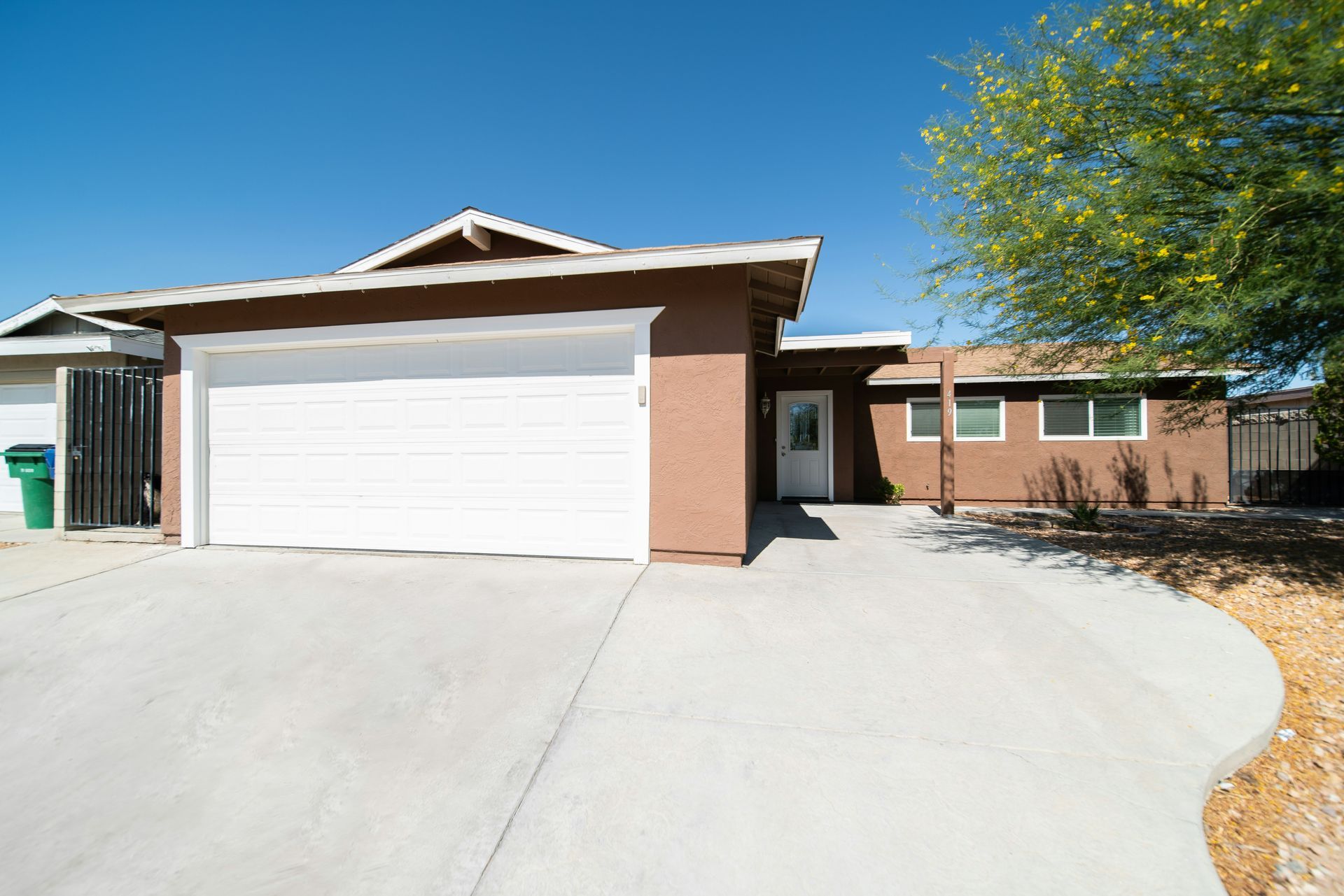 A brown single-story house with a two-car garage, a white front door, and a concrete driveway under a clear blue sky.