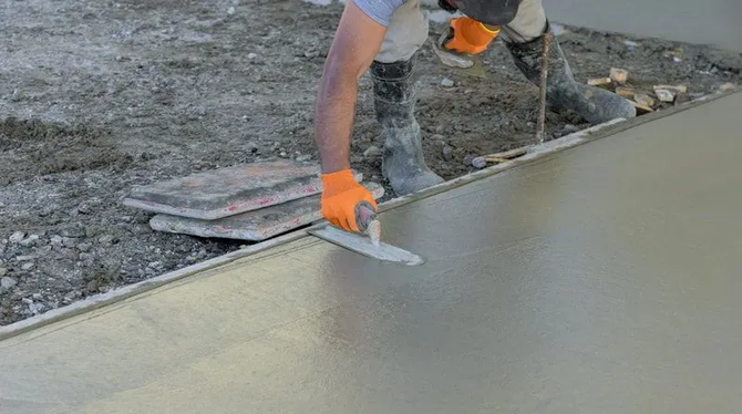 A worker wearing orange gloves uses a hand trowel to smooth freshly poured concrete in an outdoor construction area.