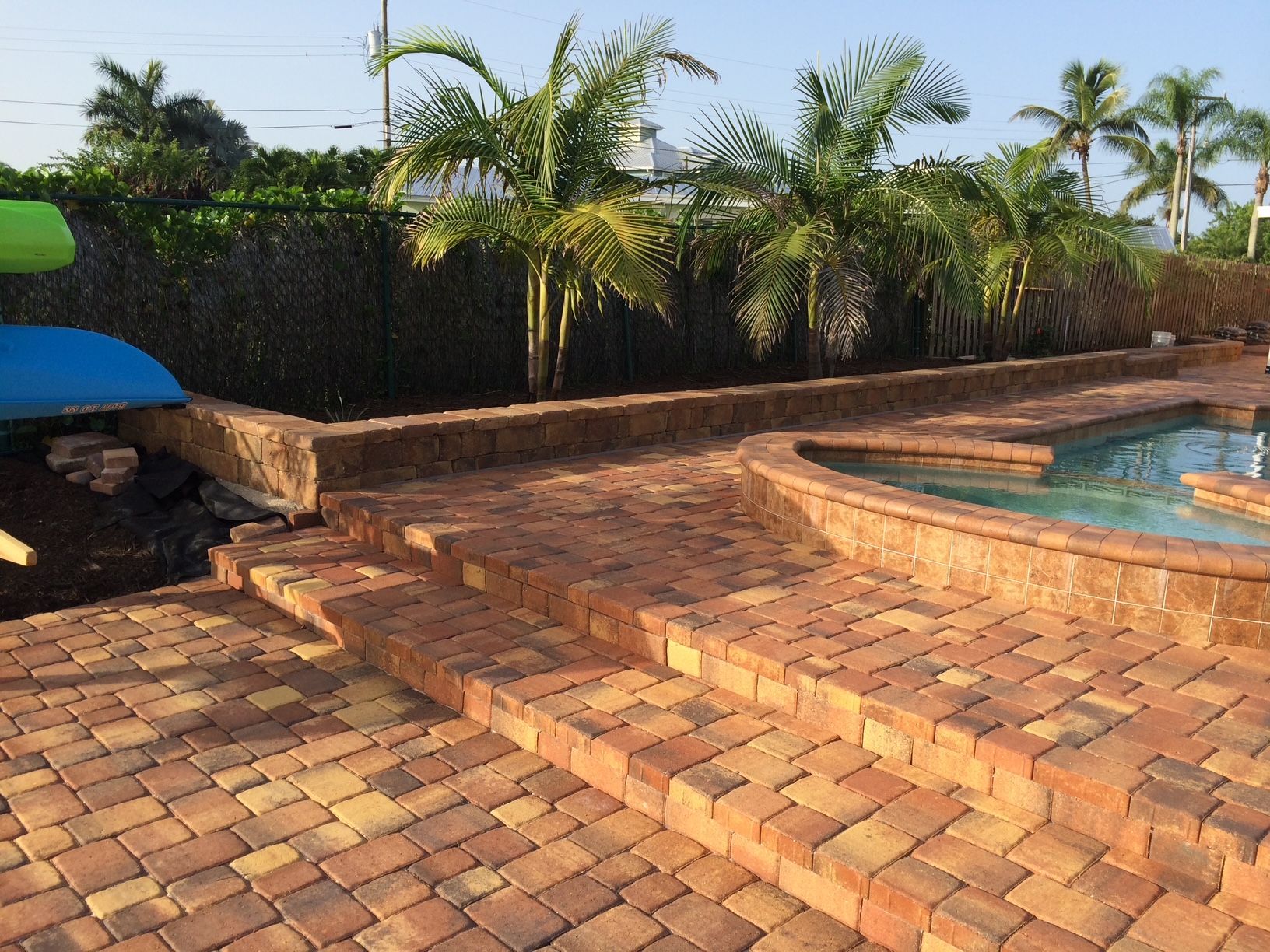 A brick patio with stairs leading up to a pool surrounded by palm trees.