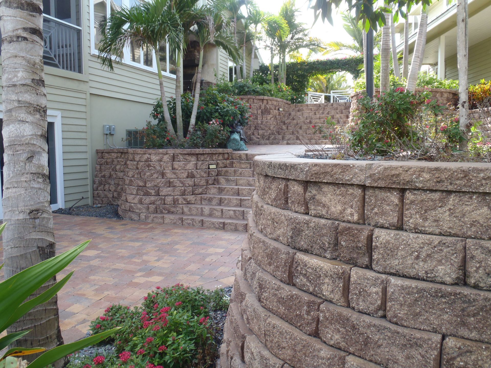 A brick wall is surrounded by flowers and trees in front of a house.