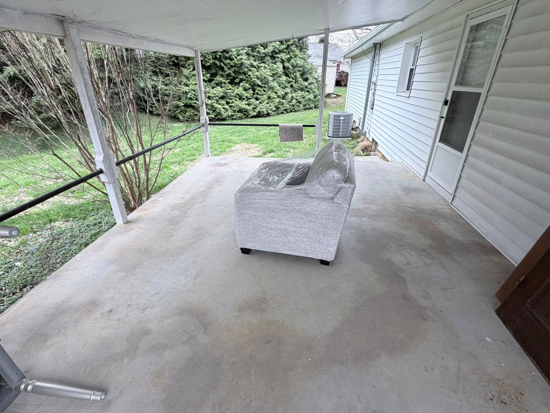 Covered concrete porch with gray loveseat and chair beside a white house and green yard.