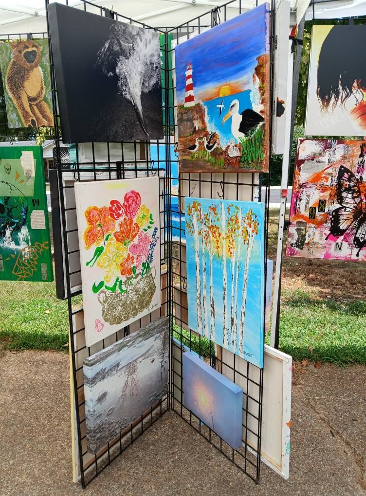 Art display with various paintings on a metal grid, outdoors. Includes landscapes and floral artwork.