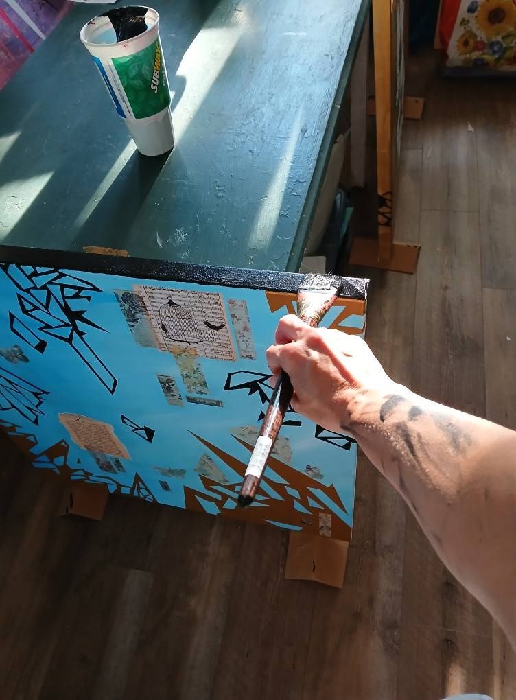 Hand paints a blue and gold artwork on a tilted table; a coffee cup sits on the green tabletop.