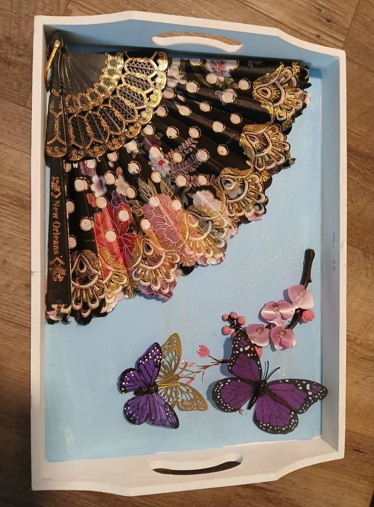 Tray with a black and gold lace fan, butterflies, and pink flowers against a light blue background.