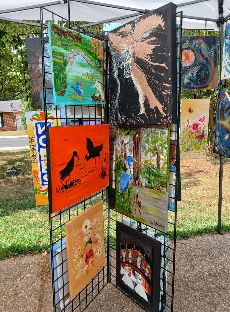 Artwork displayed on a black grid, outdoors. Includes landscape, abstract, and silhouette paintings.