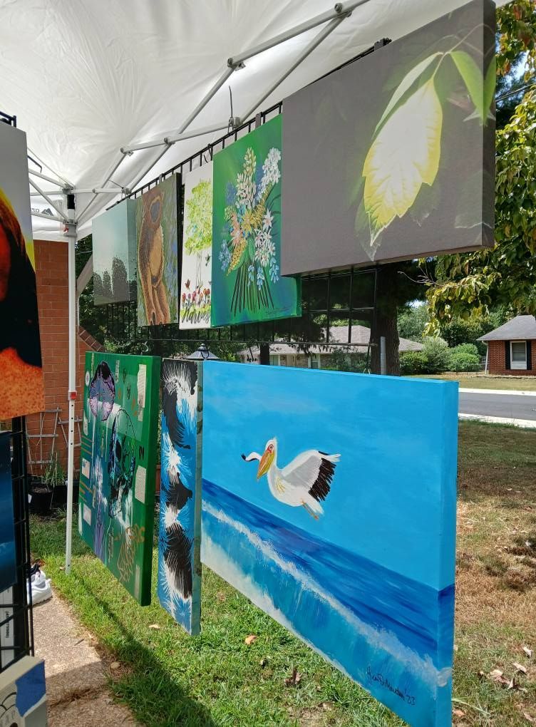 Art displayed at an outdoor art fair; paintings of birds, flowers, and landscapes hanging on a white canopy.