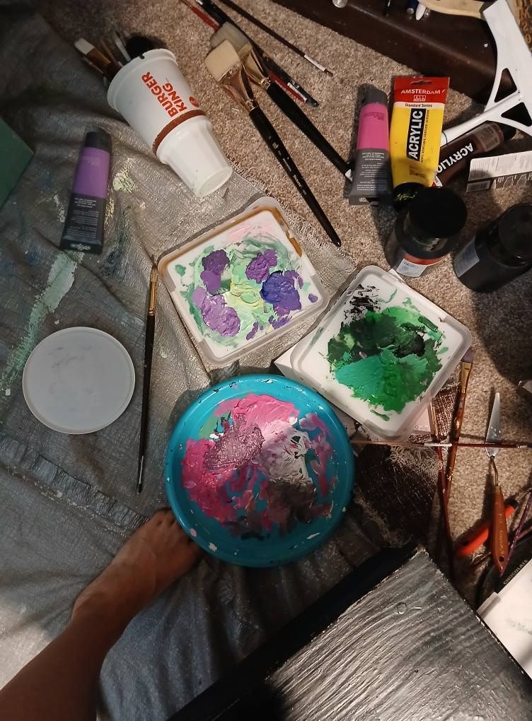 Artist's workspace with paint brushes, paints, and palette. Various colors of paint are visible on a makeshift canvas.