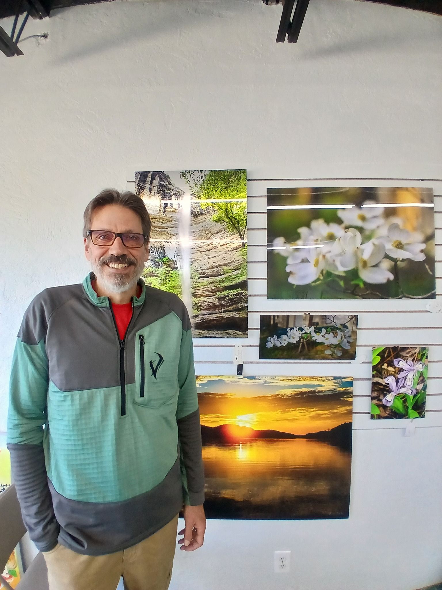 Man in front of nature photographs on a wall. He wears glasses, a green and grey jacket, and smiles.