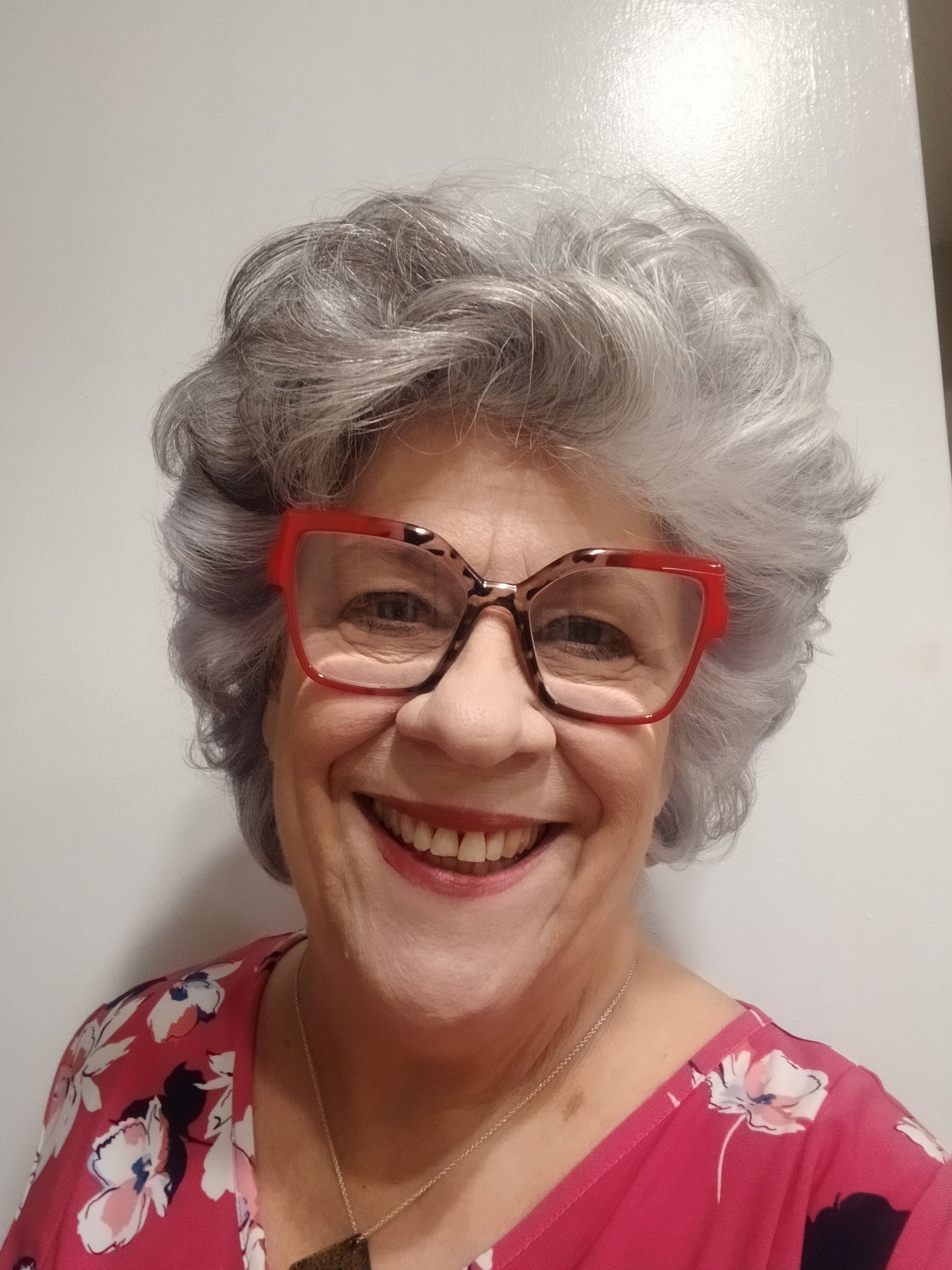 Woman with gray curly hair, wearing red-framed glasses and a pink floral top, smiling widely.