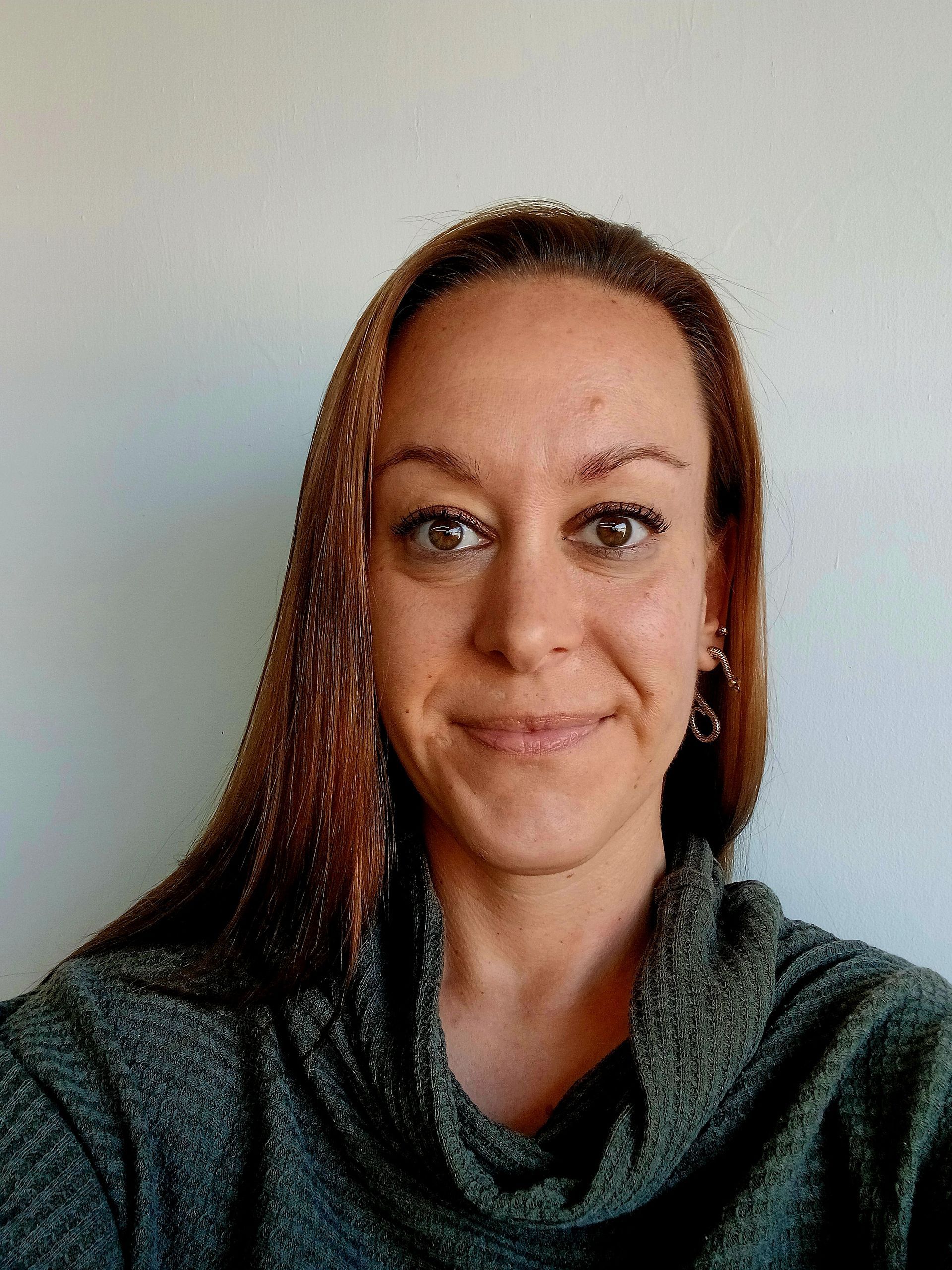 Woman with long brown hair, wearing a gray sweater, smiling in front of a white wall.