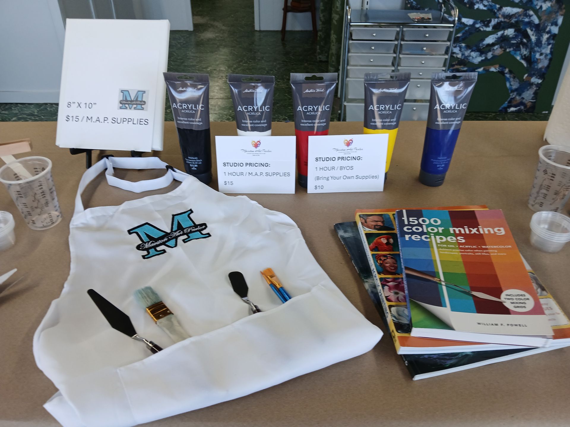 Art supplies: paint tubes, apron, mixing book, palette knives, and brushes on a table.