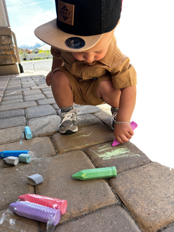 Image of a young boy aged 1 year squatting down drawing on driveway pavers with a pink piece of large chalk 