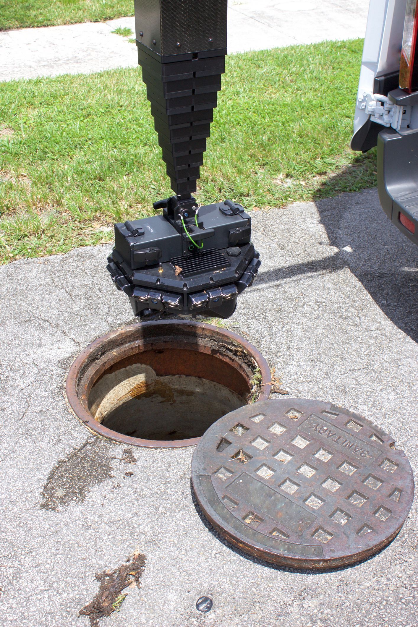 A robotic device descending into an open manhole. Manhole cover nearby. Green grass in the background.