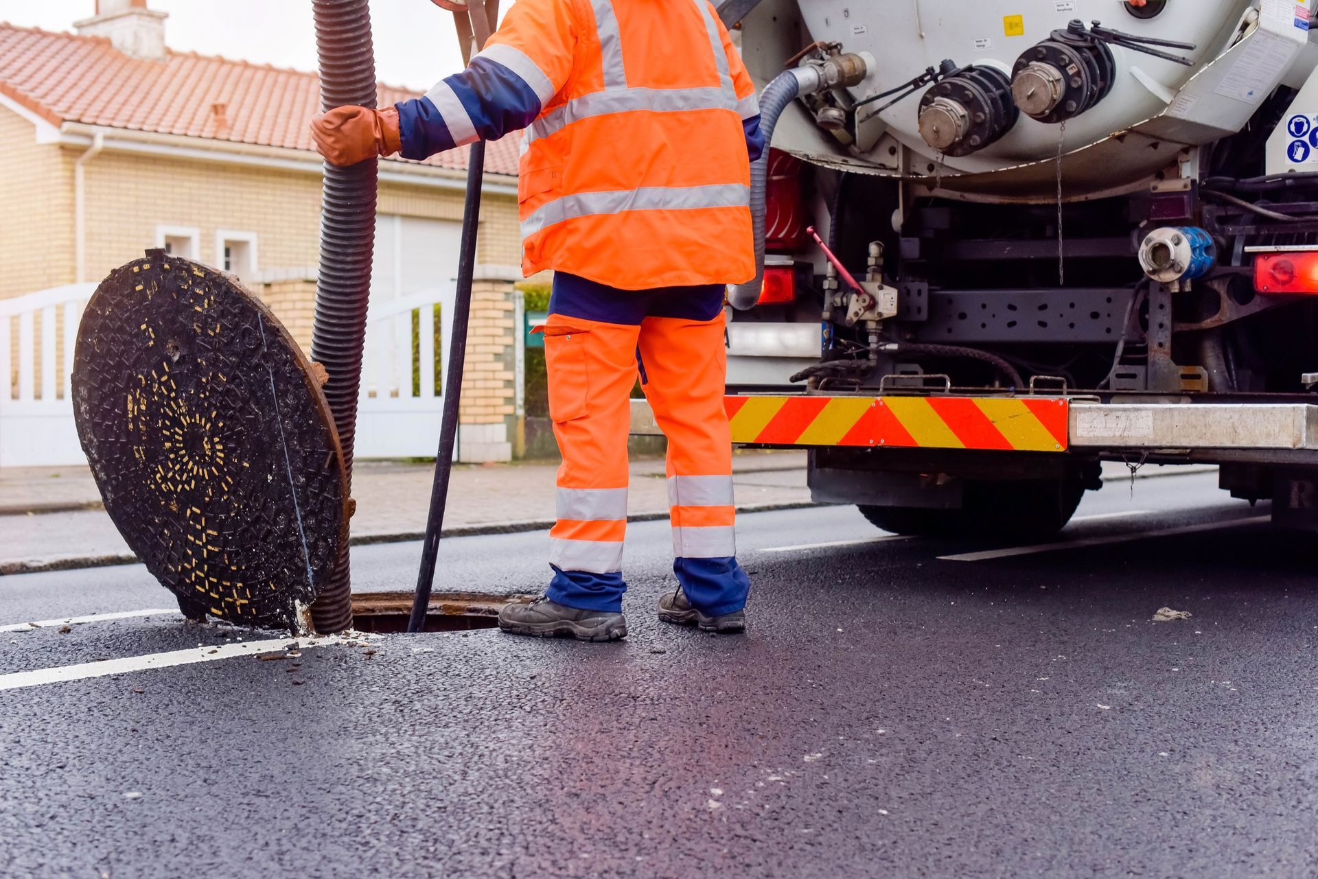 Worker in orange safety gear, hose in hand, near an open sewer drain with truck in background.