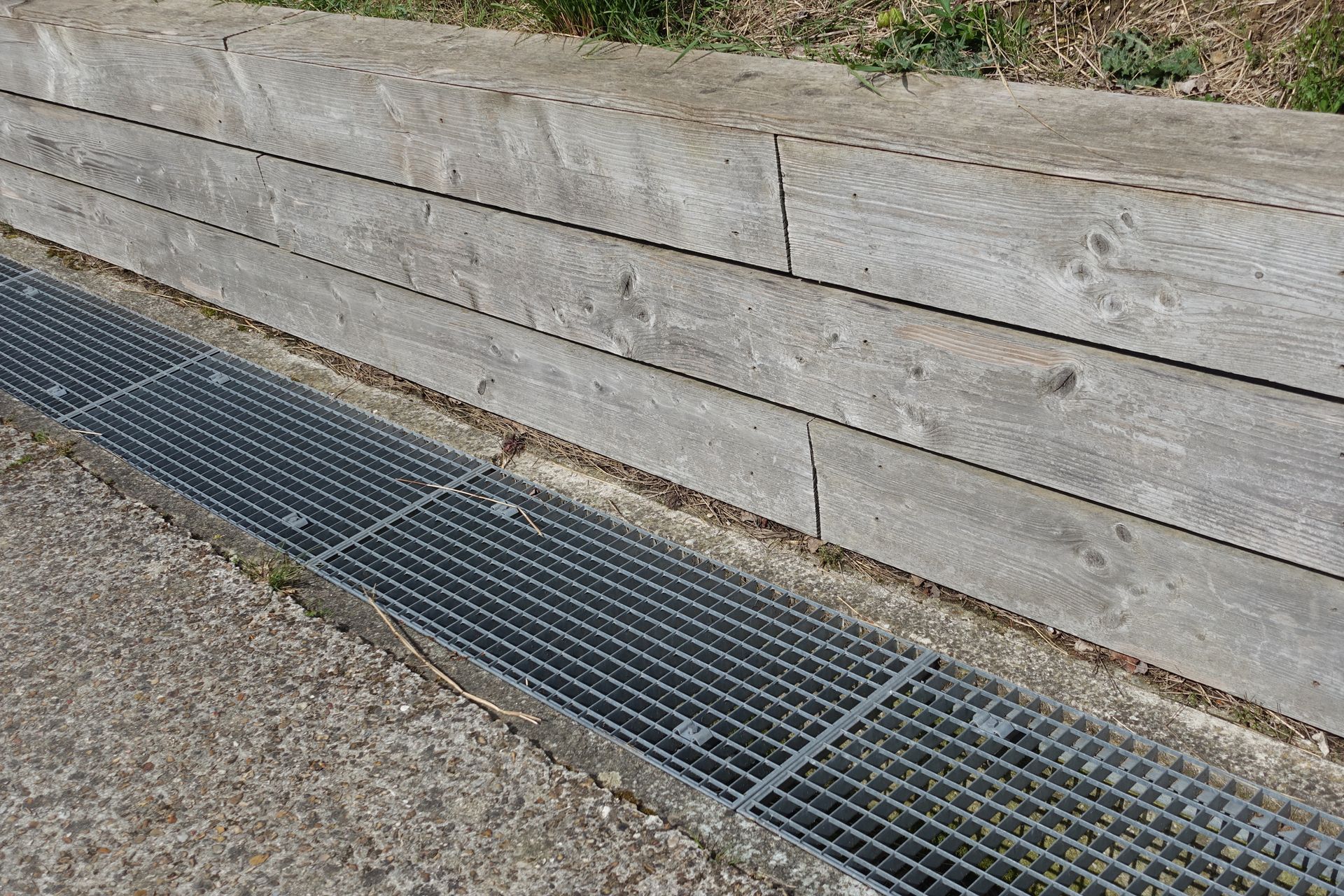 Metal grate drain next to a concrete surface and a stacked wooden wall.