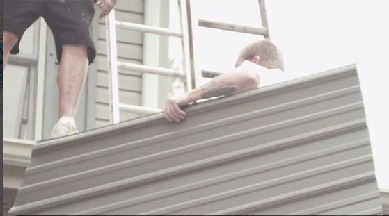 Person on roof holds sheet of metal, assisted by person on ladder.