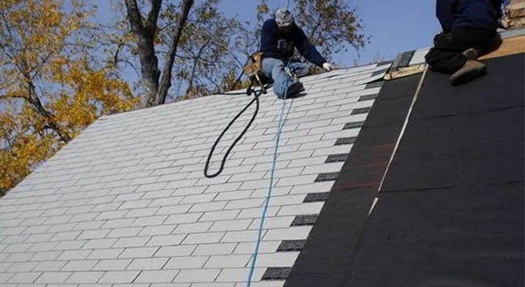 Workers on a roof installing shingles, one person is secured with a safety harness.