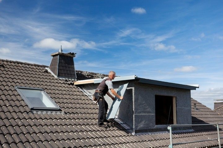 Roofer installing siding on a dormer, working on a dark tiled roof under a blue sky.