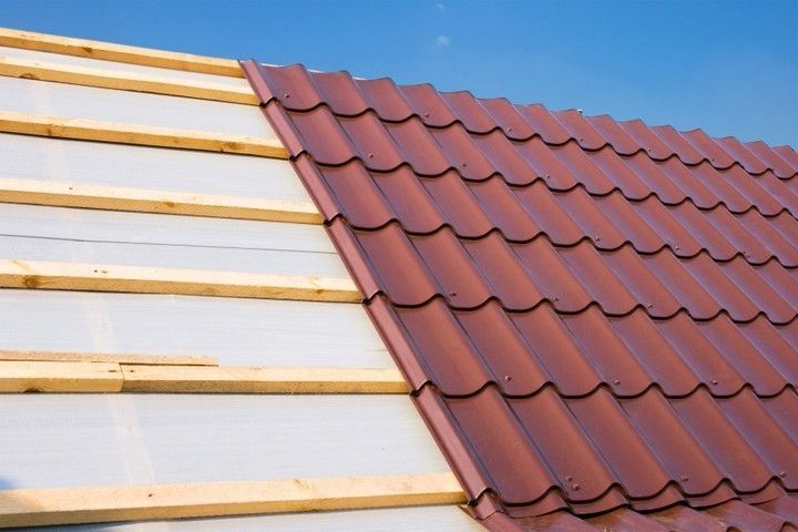Roof under construction: red metal tiles partially installed on a wooden frame against a blue sky.