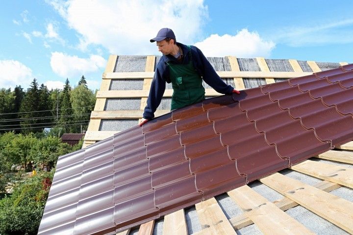 Roofer installing brown metal roofing on a wooden structure under a blue sky.