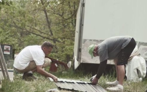 Two people installing metal roofing panels outdoors near a white trailer.
