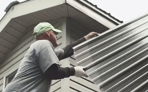 Man in green hat installs corrugated metal roofing on a house exterior.