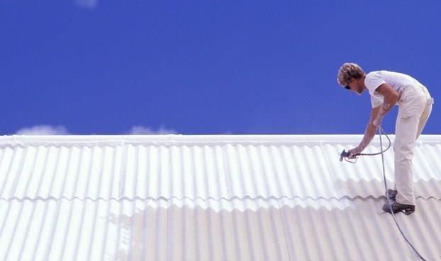 Person in white overalls spraying paint onto a white corrugated roof under a bright blue sky.