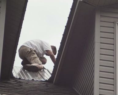 A person kneels on a metal roof, likely repairing it. The setting is outdoors.